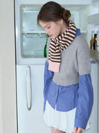 Close-up of a model wearing a layered outfit with a grey cropped sweater, blue checkered shirt, white ruffled skirt, and striped scarf, looking down.