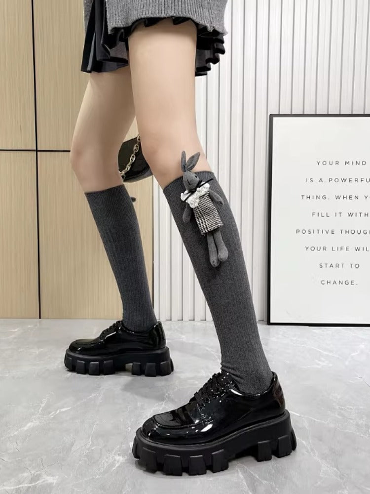 Full body shot of a person wearing grey knee-high socks with a bunny charm and black platform shoes, standing in front of a wall with a framed quote.