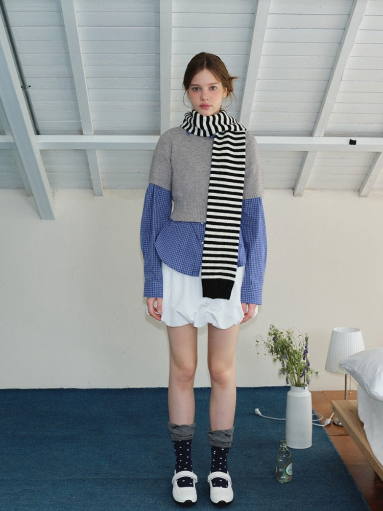 Model wearing a layered outfit with a grey cropped sweater over a blue checkered shirt, white ruffled skirt, striped scarf, and white Mary Jane shoes, standing in a bedroom setting.