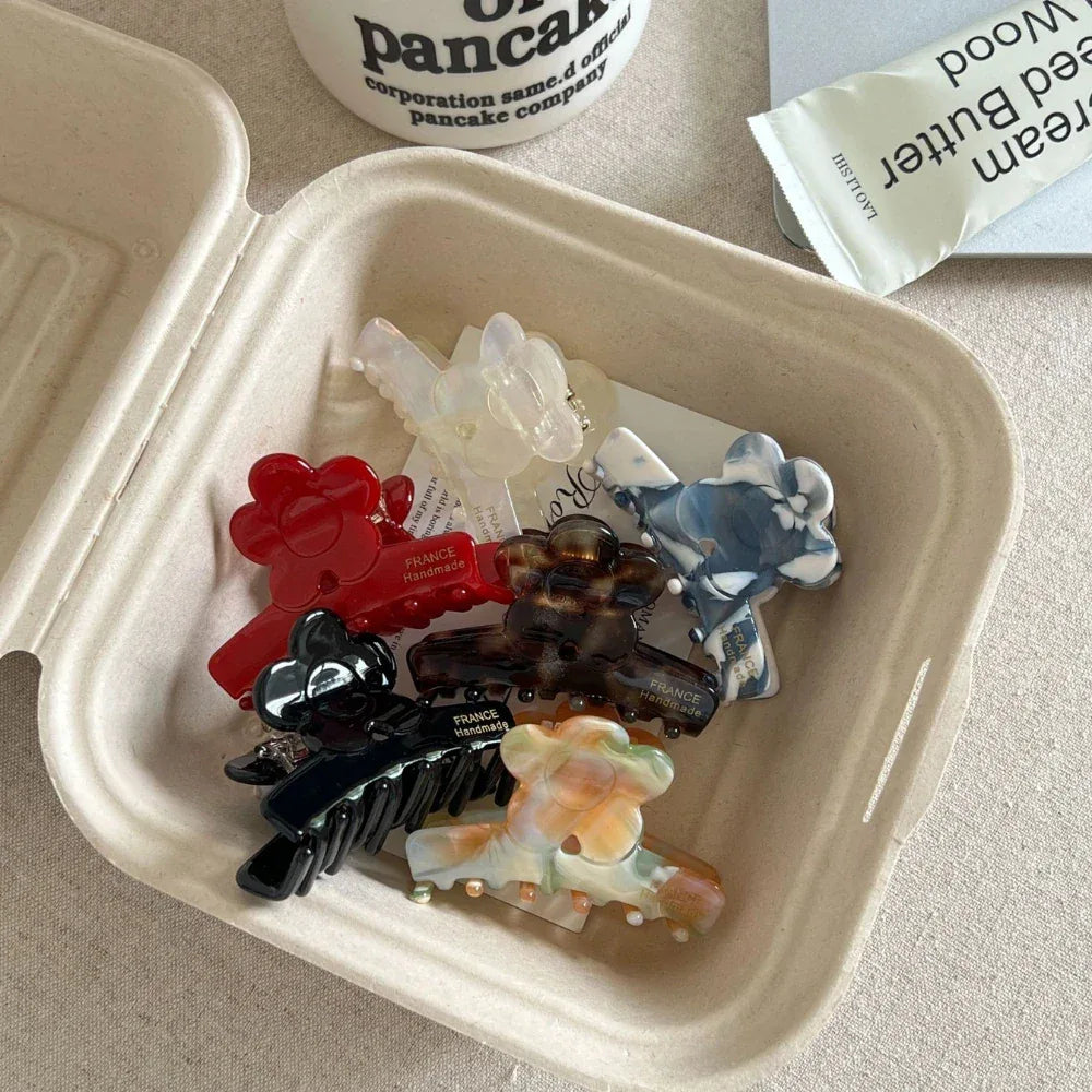 A set of flower-shaped claw clips, in colors like red, black, tortoiseshell, and marbled patterns, are neatly placed inside a small, open cardboard container.