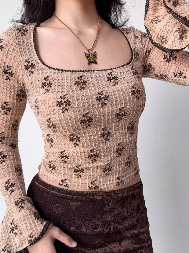 A person wearing a fitted, long-sleeved square neck top with an allover brown floral print on a beige textured knit fabric. The top features flared sleeves with dark scalloped trim. A brown velvet skirt is worn below.