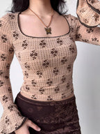 A person wearing a fitted, long-sleeved square neck top with an allover brown floral print on a beige textured knit fabric. The top features flared sleeves with dark scalloped trim. A brown velvet skirt is worn below.