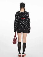 Back view of a model wearing a black zip-up hoodie with an all-over white Hello Kitty and heart graphic print, black shorts, black knee-high socks, and red sneakers. A red bow is visible on the back of the hood.