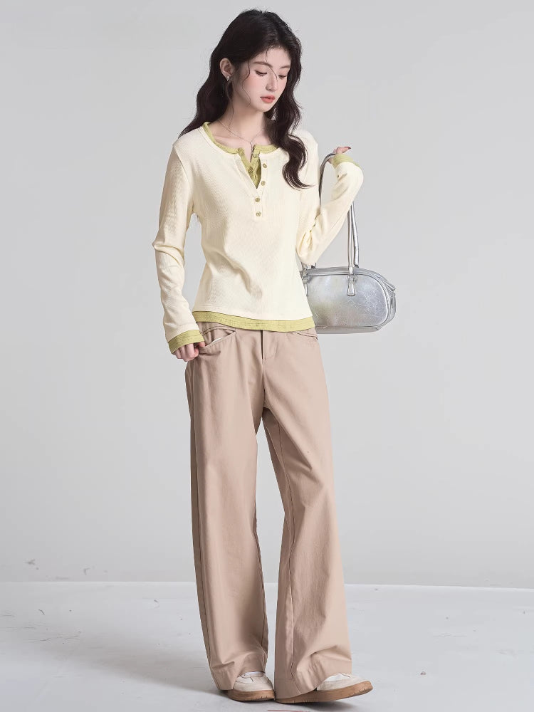 Full-body shot of a woman wearing a cream long-sleeved shirt with green trim and buttons, khaki pants, and holding a silver handbag.