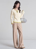 Full-body shot of a woman wearing a cream long-sleeved shirt with green trim and buttons, khaki pants, and holding a silver handbag.