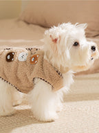 Rear view of a white dog wearing a tan fleece vest with embroidered bear appliques and a checkered trim.