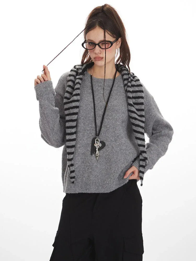 Woman wearing a grey knit sweater with a striped scarf, black cargo pants, and glasses, holding her scarf.