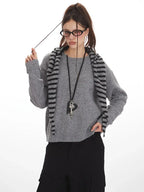 Woman wearing a grey knit sweater with a striped scarf, black cargo pants, and glasses, holding her scarf.
