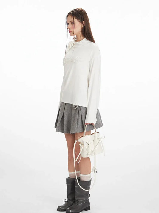 Side view of a woman wearing a white long-sleeved top with one shoulder exposed and a grey pleated skirt, holding a white handbag.