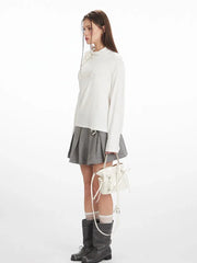 Side view of a woman wearing a white long-sleeved top with one shoulder exposed and a grey pleated skirt, holding a white handbag.