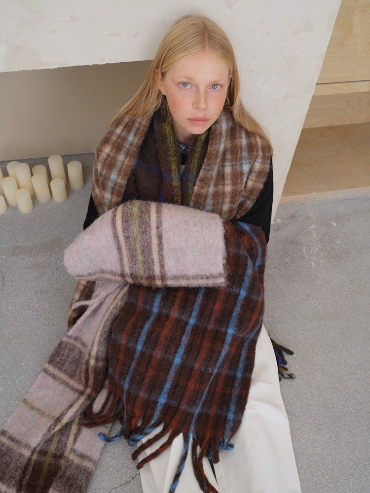 A young woman with blonde hair and freckles sits on the floor, wrapped in a large, plaid scarf. The scarf has a pattern of brown, blue, and pink squares.