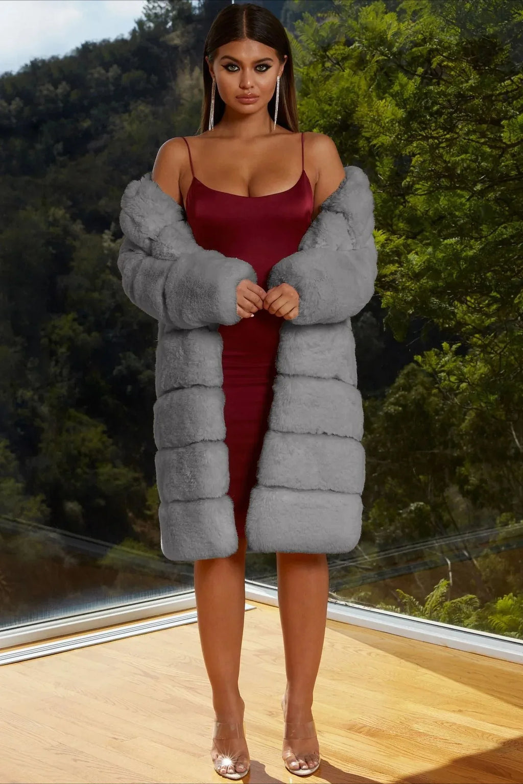 Woman wearing a gray faux fur coat over a burgundy slip dress, standing indoors with a scenic view.