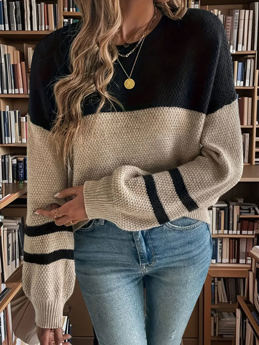 Woman wearing a black and tan color-blocked sweater with striped sleeves and blue jeans, standing in front of a bookshelf.