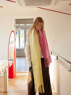 Woman wearing a two-tone yellow and pink fringed scarf draped over a white crochet top and brown wide-leg pants, looking down while leaning on a railing.