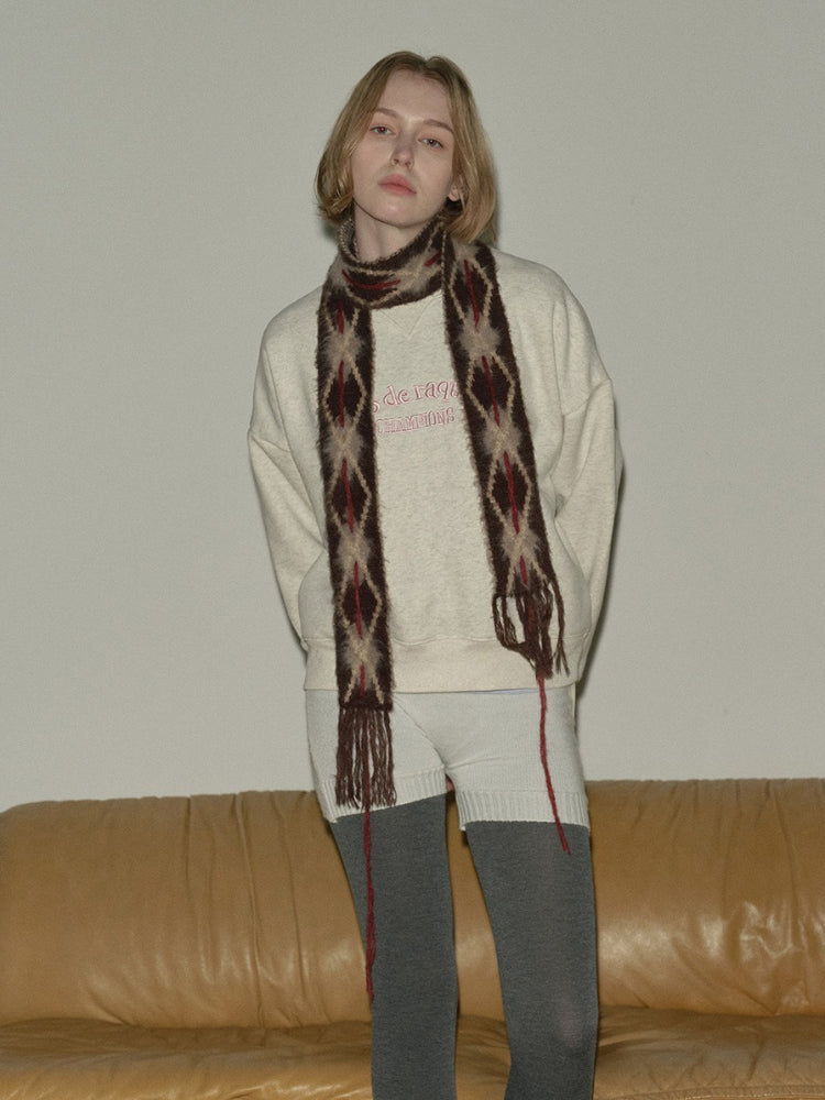 Model standing in front of a couch wearing a cream sweatshirt and argyle scarf