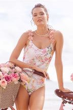 Woman in a floral print one-piece swimsuit with ruffled straps, posing with a bicycle and a basket of flowers.