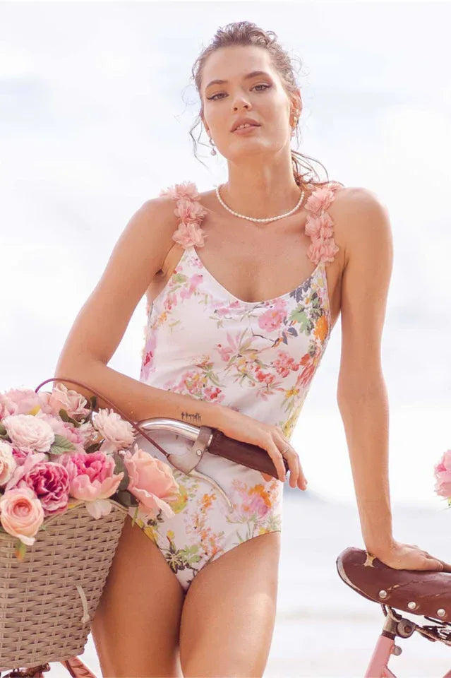 Woman in a floral print one-piece swimsuit with ruffled straps, posing with a bicycle and a basket of flowers.