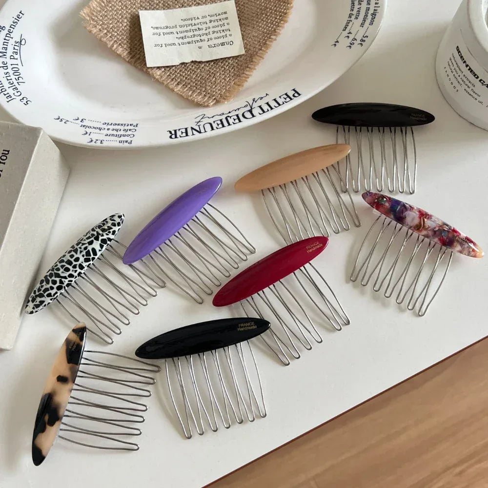 Several decorative hair combs with oval tops in different colors and patterns are displayed on a white surface, with some fabric and decorative items in the background.