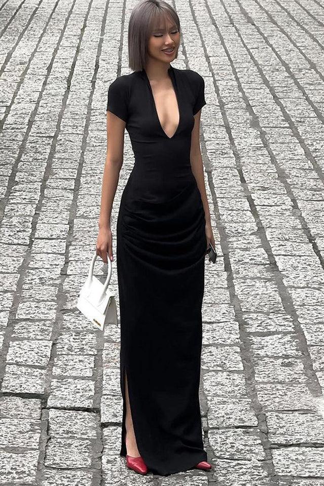 Full body shot of a model wearing a black long dress with a deep V-neckline and ruching on the skirt, holding a white bag with fringe details, walking on a cobblestone street.