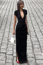 Full body shot of a model wearing a black long dress with a deep V-neckline and ruching on the skirt, holding a white bag with fringe details, walking on a cobblestone street.