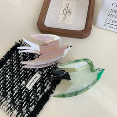 Two acrylic bird hair clips, one pearlescent pink and white, the other green and white, displayed on a textured black and white cloth.