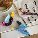 Three colorful acrylic resin bird brooches displayed on a white surface with a piece of bread and a cloth napkin.