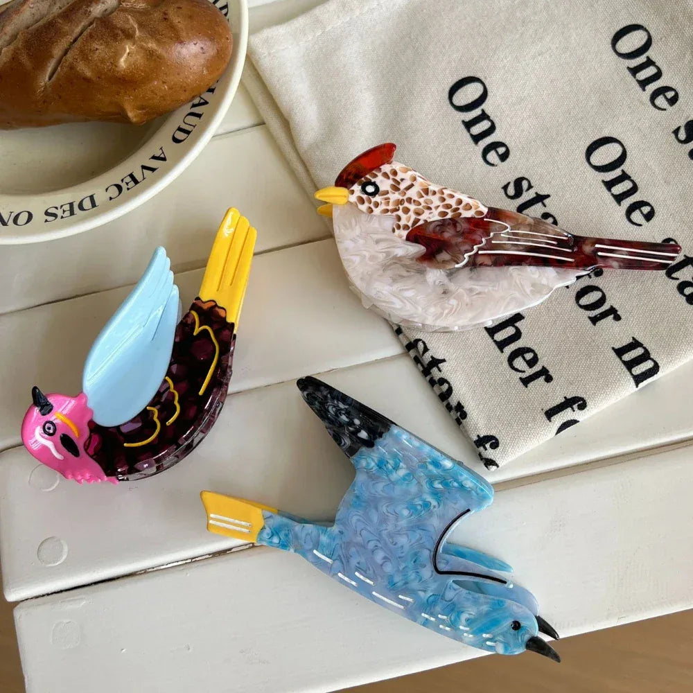 Three colorful acrylic resin bird brooches displayed on a white surface with a piece of bread and a cloth napkin.