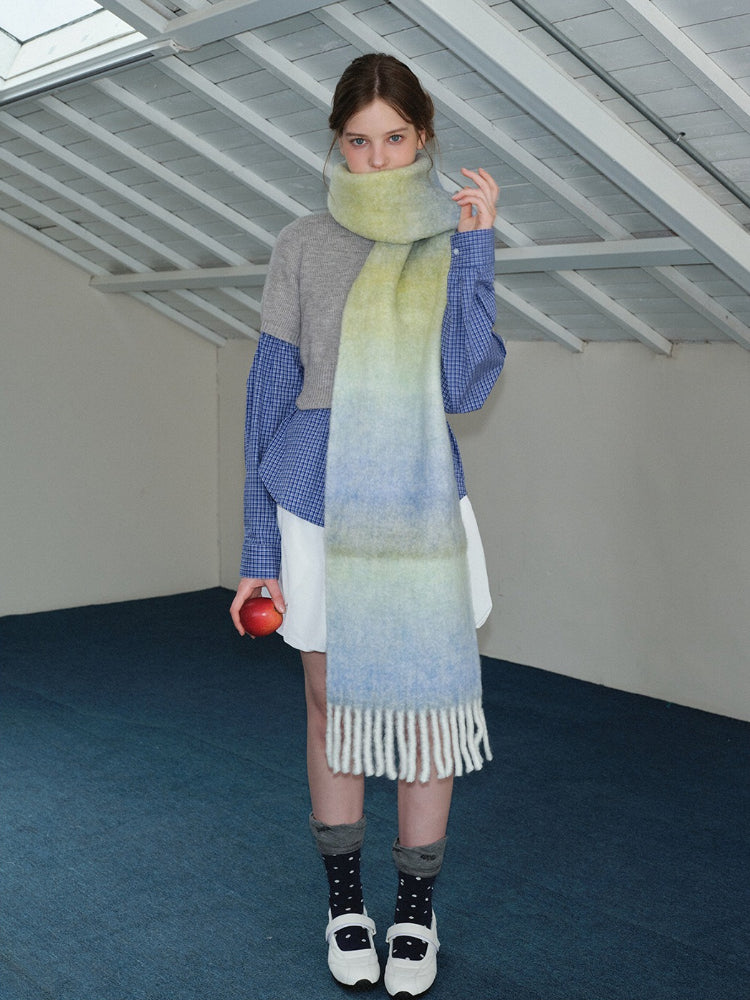 Woman wearing an oversized ombre scarf with a grey sweater, blue plaid shirt, and white shorts, holding a red apple.