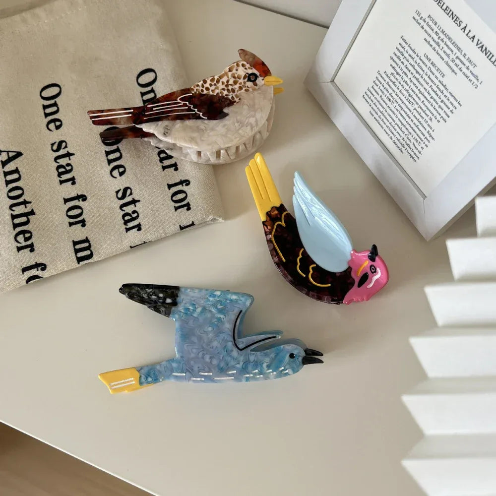 An overhead shot of three acrylic resin bird brooches arranged on a white wooden surface, with a linen cloth and part of a box visible.