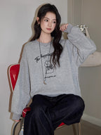 Woman sitting on a red chair wearing a grey crew neck sweatshirt with a graphic print and dark wash jeans