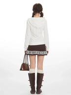 Back view of a woman wearing a white zip-up hoodie, a brown patterned skirt, white socks, and brown boots, holding a brown handbag.