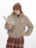 Model wearing a beige fuzzy bomber jacket and red plaid pants, reading a book.