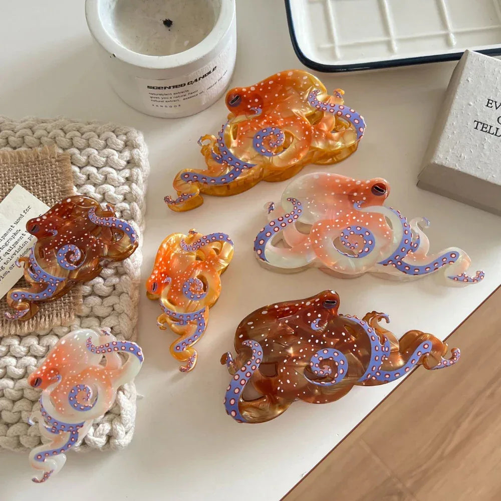 A flat lay of several octopus-shaped hair clips in different translucent colors and sizes, featuring white polka dots and blue tentacle details, displayed on a white table with various accessories.