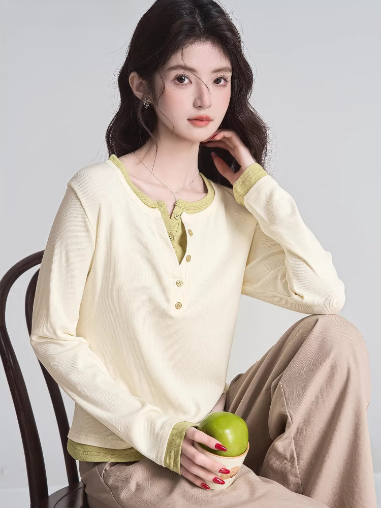 Woman sitting on a chair wearing a cream long-sleeved shirt with green trim and buttons, holding an apple.