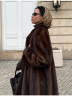 Side profile of a woman wearing a long, brown mink fur coat, black turtleneck, black leggings, black woven handbag, and brown pointed-toe heels.