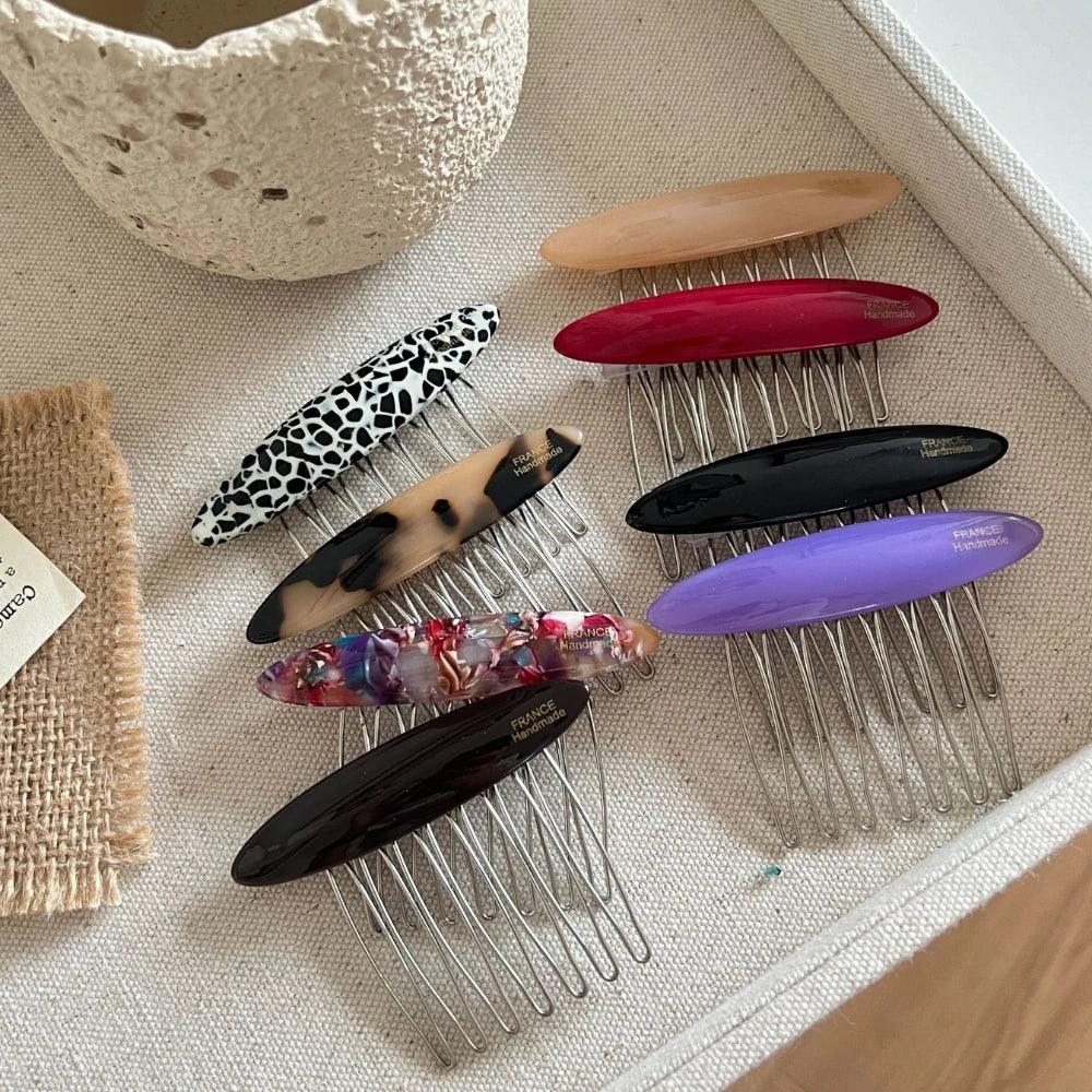 A collection of hair combs with decorative oval tops in various colors and patterns, including animal print, tortoiseshell, solid colors, and marbled designs, arranged on a textured surface.