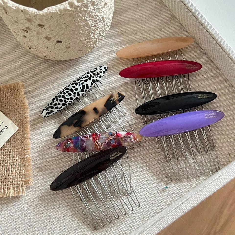 A collection of hair combs with decorative oval tops in various colors and patterns, including animal print, tortoiseshell, solid colors, and marbled designs, arranged on a textured surface. Secondary image