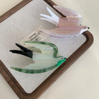 Two acrylic bird hair clips, one pearlescent pink and white, the other green and white, placed on a wooden tray with a linen insert.