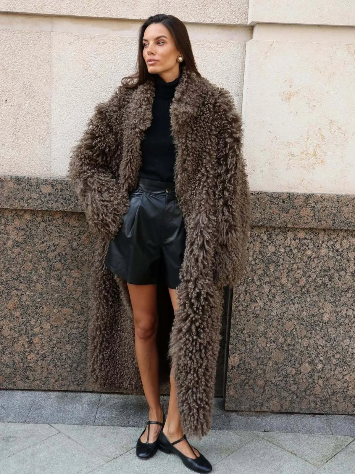 Woman wearing a long, fluffy brown faux fur coat over a black turtleneck and black leather shorts, standing against a granite-patterned wall. Main image