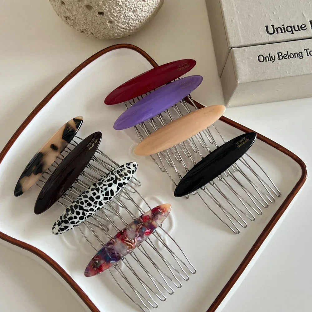 A white ceramic dish holds several hair combs with decorative oval tops in various colors and patterns, showcasing their designs and textures.