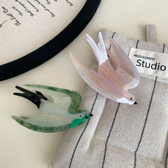 Two acrylic bird hair clips, one pearlescent pink and white, the other green and white, placed on a grey striped cloth with a label that says "MEZO ELEMENT Studio".