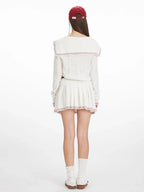 Back view of a model wearing a white knit sweater with a sailor collar and red trim, paired with a white pleated skirt and white socks.