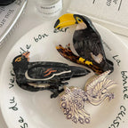 A close-up of three decorative bird hair clips on a plate, showcasing a toucan, a crow, and a peacock design.
