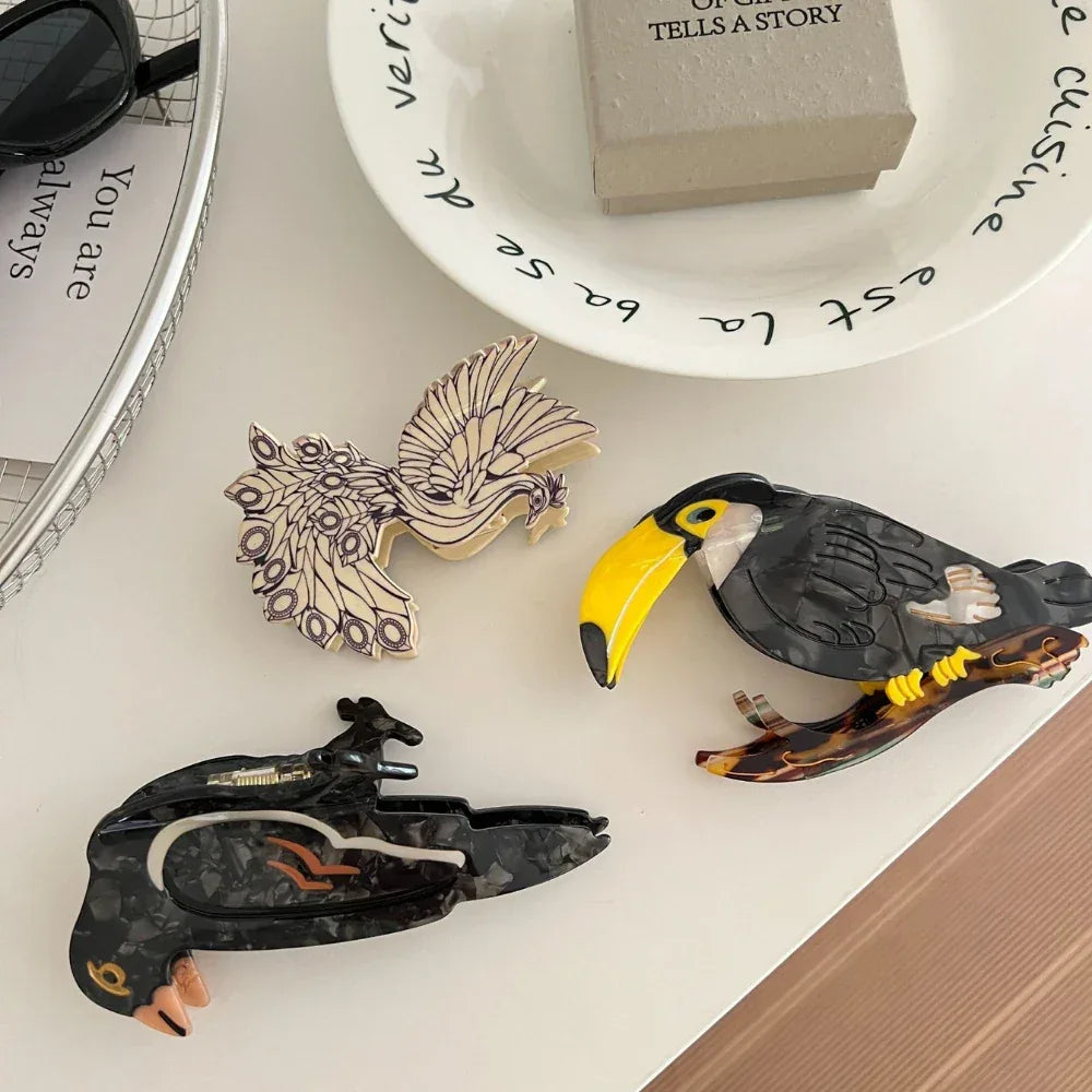 Three decorative bird hair clips - a toucan, a crow, and a peacock - scattered on a white surface with other decorative items.