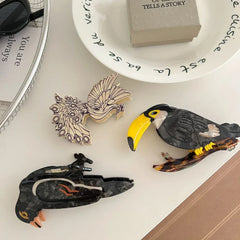 Three decorative bird hair clips - a toucan, a crow, and a peacock - scattered on a white surface with other decorative items.
