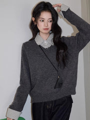Woman standing with her hand on her head, wearing a grey v-neck sweater over a checkered collared shirt, dark wash wide leg jeans, and a black crossbody pouch.