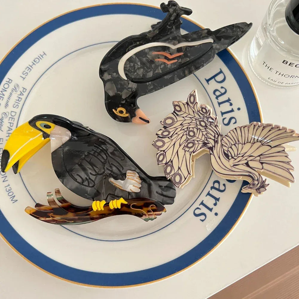 A collection of three decorative bird hair clips, including a toucan, a crow, and a peacock, arranged on a plate.