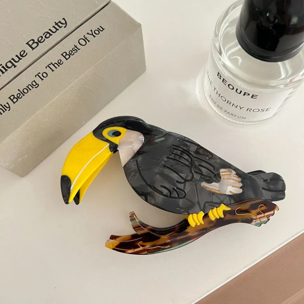 A decorative acrylic toucan hair clip with a branch accent, placed next to a perfume bottle and a box.