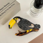 A decorative acrylic toucan hair clip with a branch accent, placed next to a perfume bottle and a box.