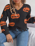 Close-up of a woman wearing a black long-sleeve v-neck cropped knit sweater with an all-over orange pumpkin print and a distressed hem, sitting on a couch.